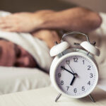sleepy-young-man-covering-ears-with-pillow-as-he-looks-alarm-clock-bed-sleeping-man-disturbed-by-alarm-clock-early-morning-frustrated-man-listening-his-alarm-clock-while-relaxing-his-bed